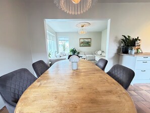 Ein Essbereich mit Holztisch, vier Stühlen, einer Chandelierleuchtre und Blick in ein Wohnzimmer mit Sofa und Gemälde.