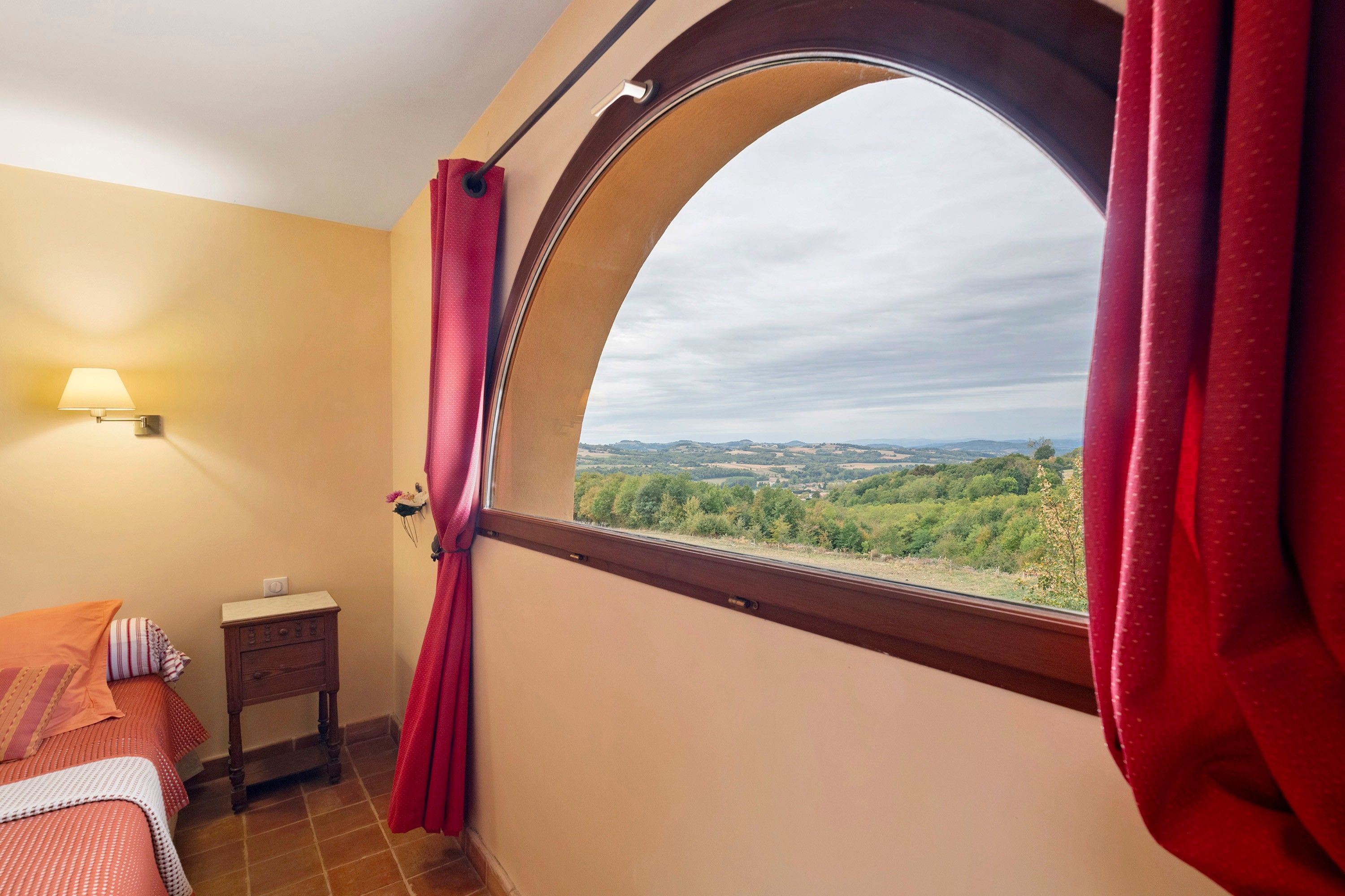 A bedroom with a curved window, a bedside table, a lamp, and a view of a landscape.