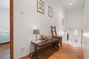 A room with a bed, a desk with a lamp, a chair, and a wall-mounted shelf.