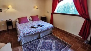 Une chambre à coucher avec un lit, deux tables de chevet, un lustre, une fenêtre avec des rideaux rouges et un tapis à motifs.