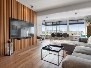 A modern living room with a flat-screen TV, wooden panel wall, large windows, a dining area, and a sofa. - Modern and family friendly house in Garðabær (Garðabær)