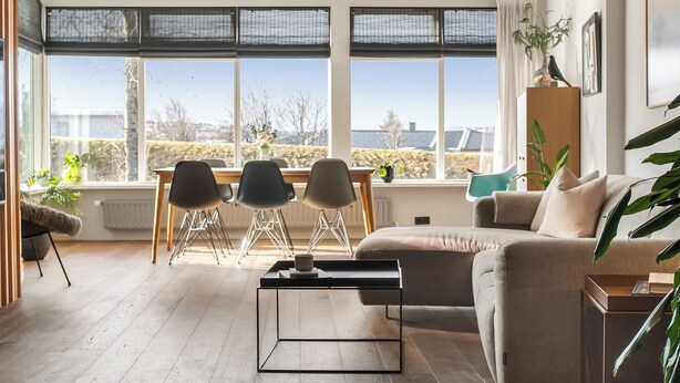 Ett modernt vardagsrum med en soffa, ett soffbord och en matplats med stolar och ett bord. Stora fönster ger utsikt över omgivningen.