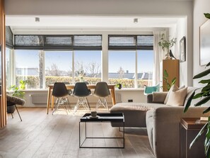 A modern living room with a sofa, a coffee table, and a dining area with chairs and a table. Large windows provide a view of the outside. - Modern and family friendly house in Garðabær (Garðabær)