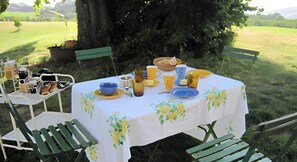 Un espace de restauration en plein air avec une table dressée pour deux personnes, entourée de verdure et d’un grand arbre.