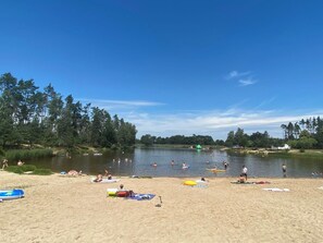 Ein Strand mit badenden und sonnenden Personen, ein See und Bäume im Hintergrund.