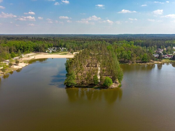 Un lago con una pequeña isla, rodeado de un bosque denso y algunos edificios.
