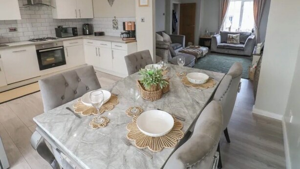 Una cocina moderna con armarios blancos, una mesa de comedor de mármol y vistas a una sala de estar con un sofá y una ventana.