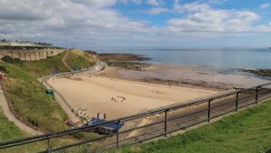 Una playa costera con un paseo marítimo cercado, una zona arenosa y una costa distante.