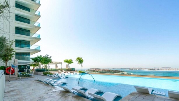 Piscina moderna de hotel con vista al mar, sillones de descanso y un edificio con balcones.