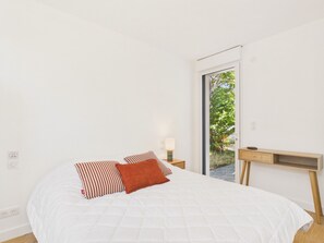 Ein Schlafzimmer mit Bett, Kissen, einem Nachttisch, einer Lampe und Blick auf Grün hinter einer Glastür.