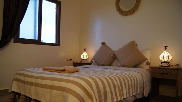 Une chambre à coucher avec un lit, deux lampes de chevet, une fenêtre, un miroir mural et une petite table de chevet en bois.