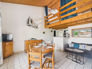 Ein Essbereich mit Holztisch und -stühlen, ein Küchenbereich mit Arbeitsplatte und Geräten sowie eine Sitzecke mit Kissen.