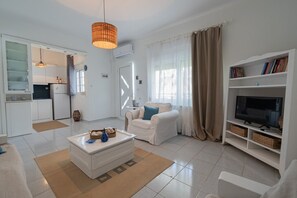 Una sala de estar moderna con un sofá blanco, una mesa de centro, un mueble para la televisión y una zona de cocina con armarios blancos y una lámpara colgante.