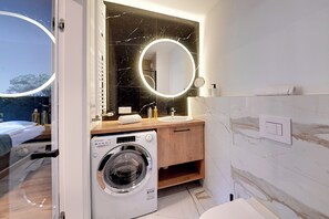 Ein modernes Badezimmer mit integrierter Waschmaschine, einem runden Spiegel mit Beleuchtung, einem Holz-Waschtisch und einer Marmorwand.