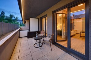 Ein Balkon mit Tisch und Stühlen, eine Schiebetür zu einem Zimmer und im Hintergrund Bäume sowie Gebäude.