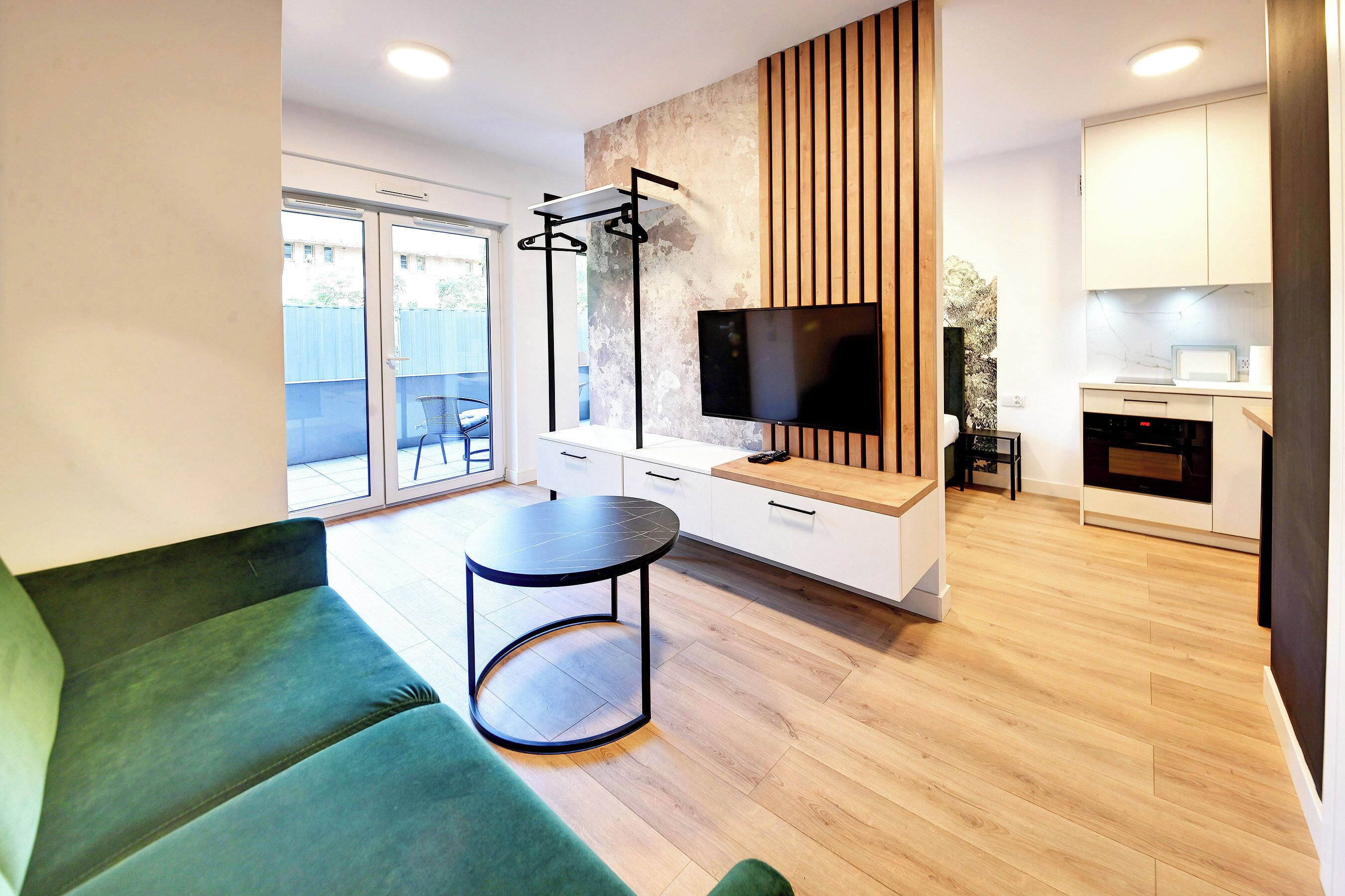 Una sala moderna con un sofá verde, una mesa redonda, un televisor y una zona de cocina con armarios blancos y un microondas.