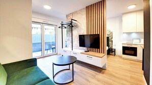 Una sala moderna con un sofá verde, una mesa redonda, un televisor y una zona de cocina con armarios blancos y un microondas.
