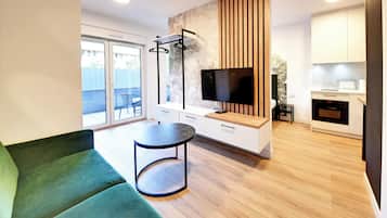 Una sala moderna con un sofá verde, una mesa redonda, un televisor y una zona de cocina con armarios blancos y un microondas.
