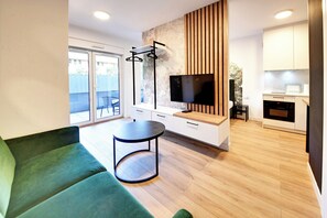 Una sala moderna con un sofá verde, una mesa redonda, un televisor y una zona de cocina con armarios blancos y un microondas.