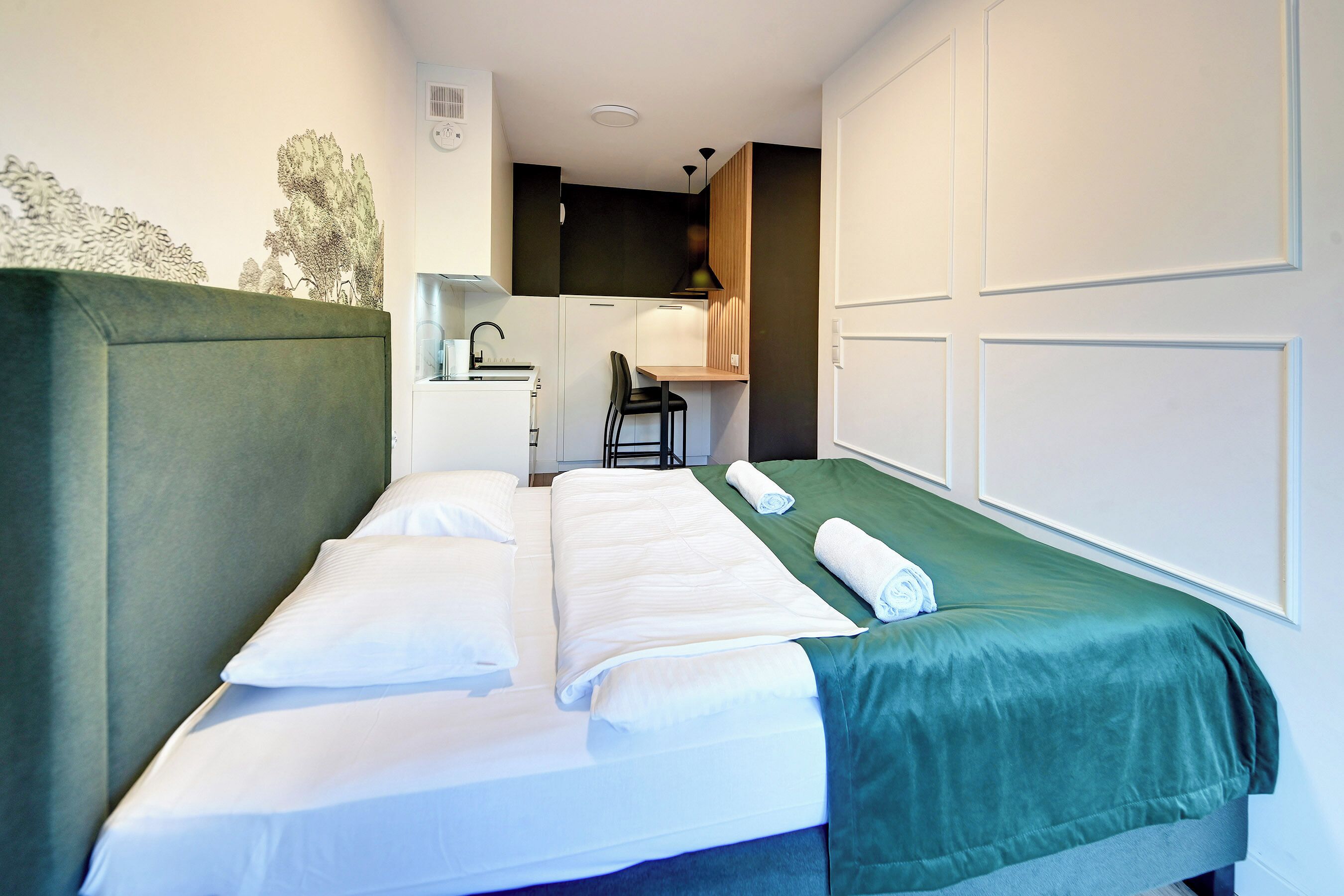 A single bed with white bedding, a green throw, a desk with a chair, and a kitchenette in a compact room.