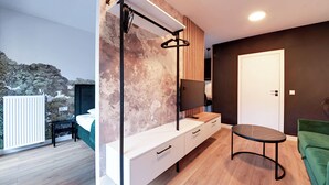 Una habitación moderna con cama, escritorio, televisor, sofá verde y vistas a árboles.