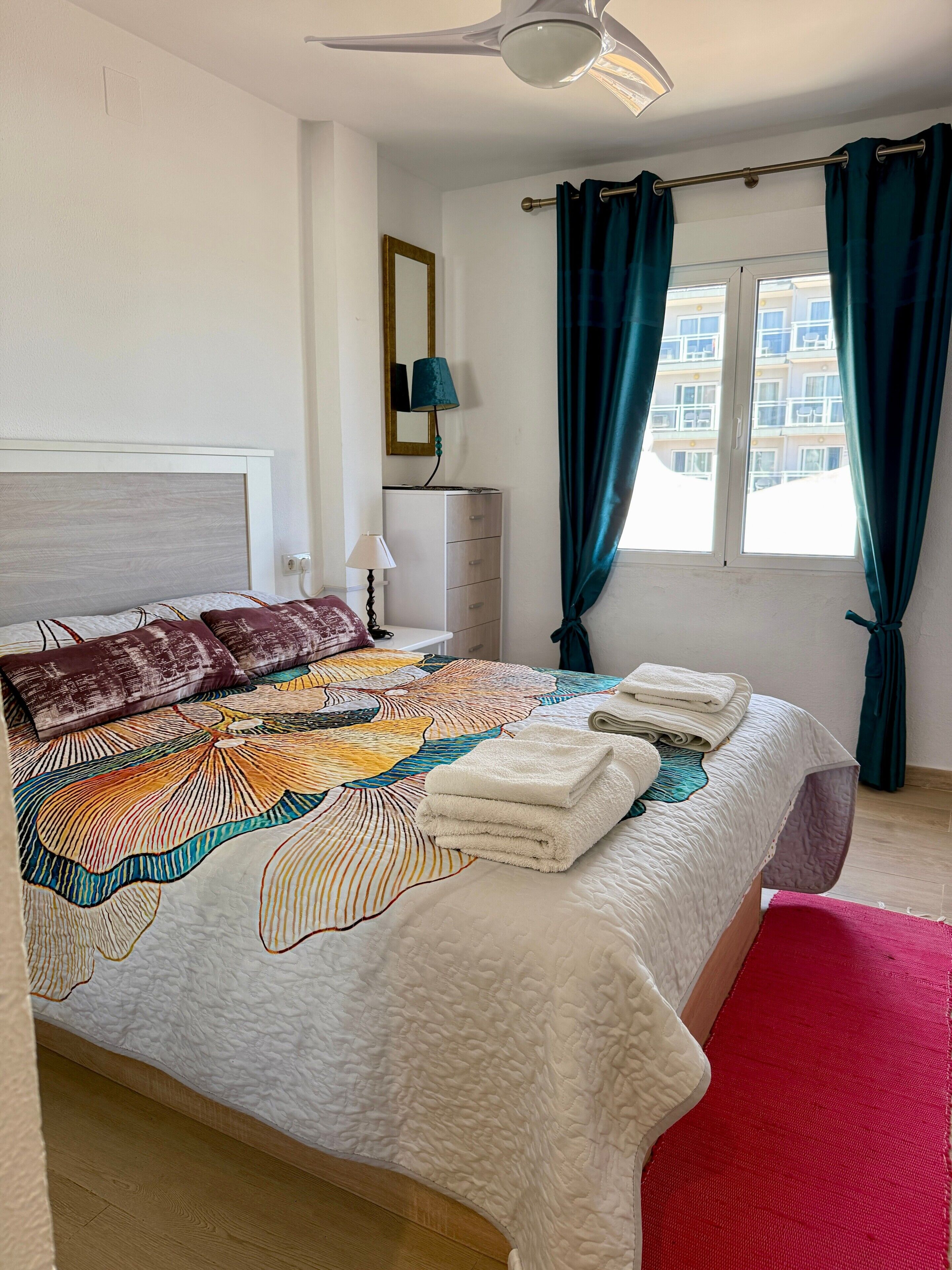 Een slaapkamer met een bed, een kaptafel, een spiegel, een plafondventilator en een raam met gordijnen.
