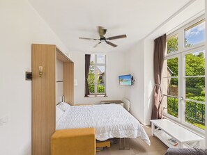 A bedroom with a bed, a desk, a chair, a ceiling fan, and a window with a view of greenery. - Apartment in a castle with pool – 4 guests (Baden)