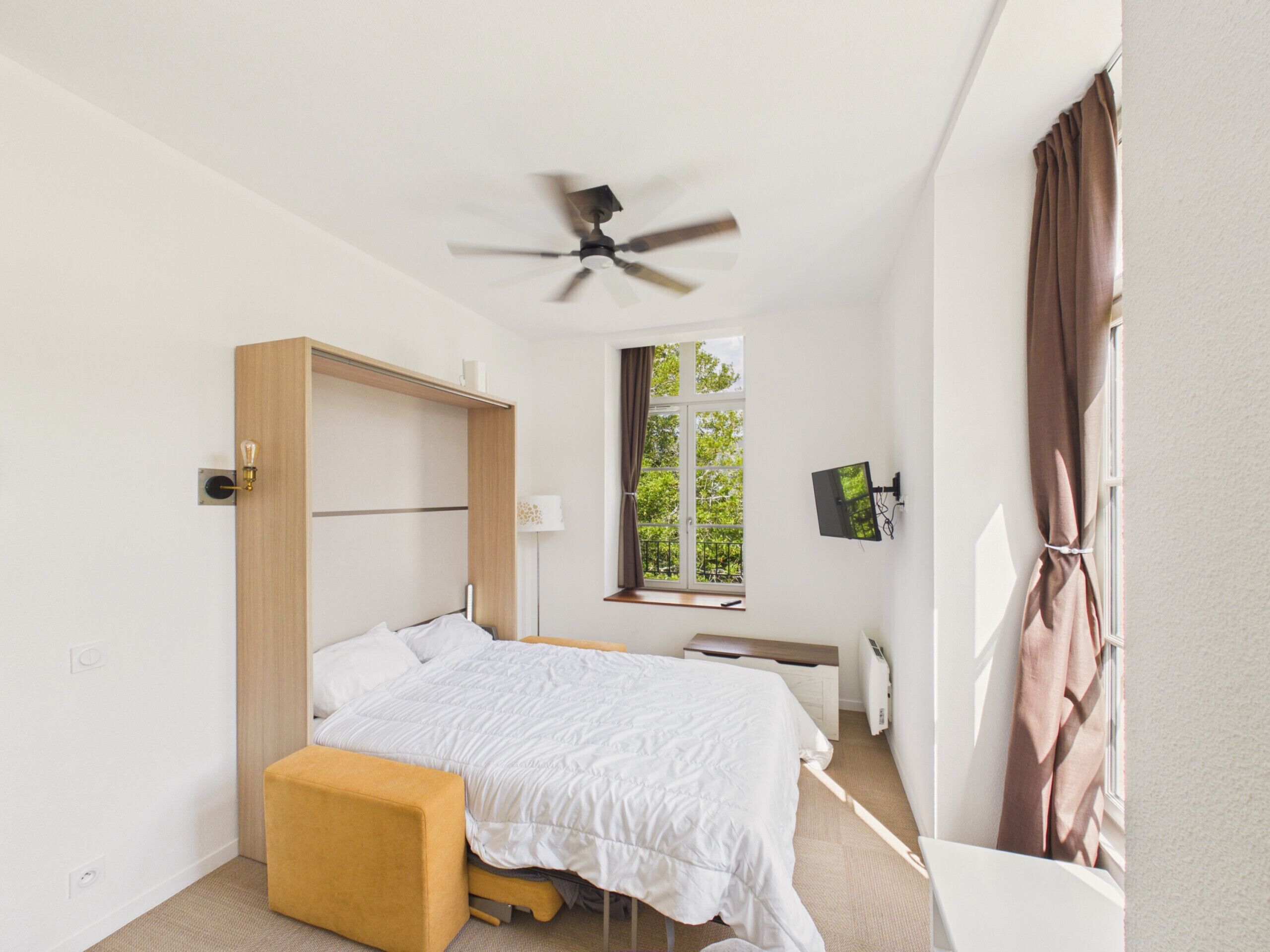 Une chambre à coucher comprenant un lit, un ventilateur de plafond, une fenêtre avec des rideaux, un bureau et une chaise.