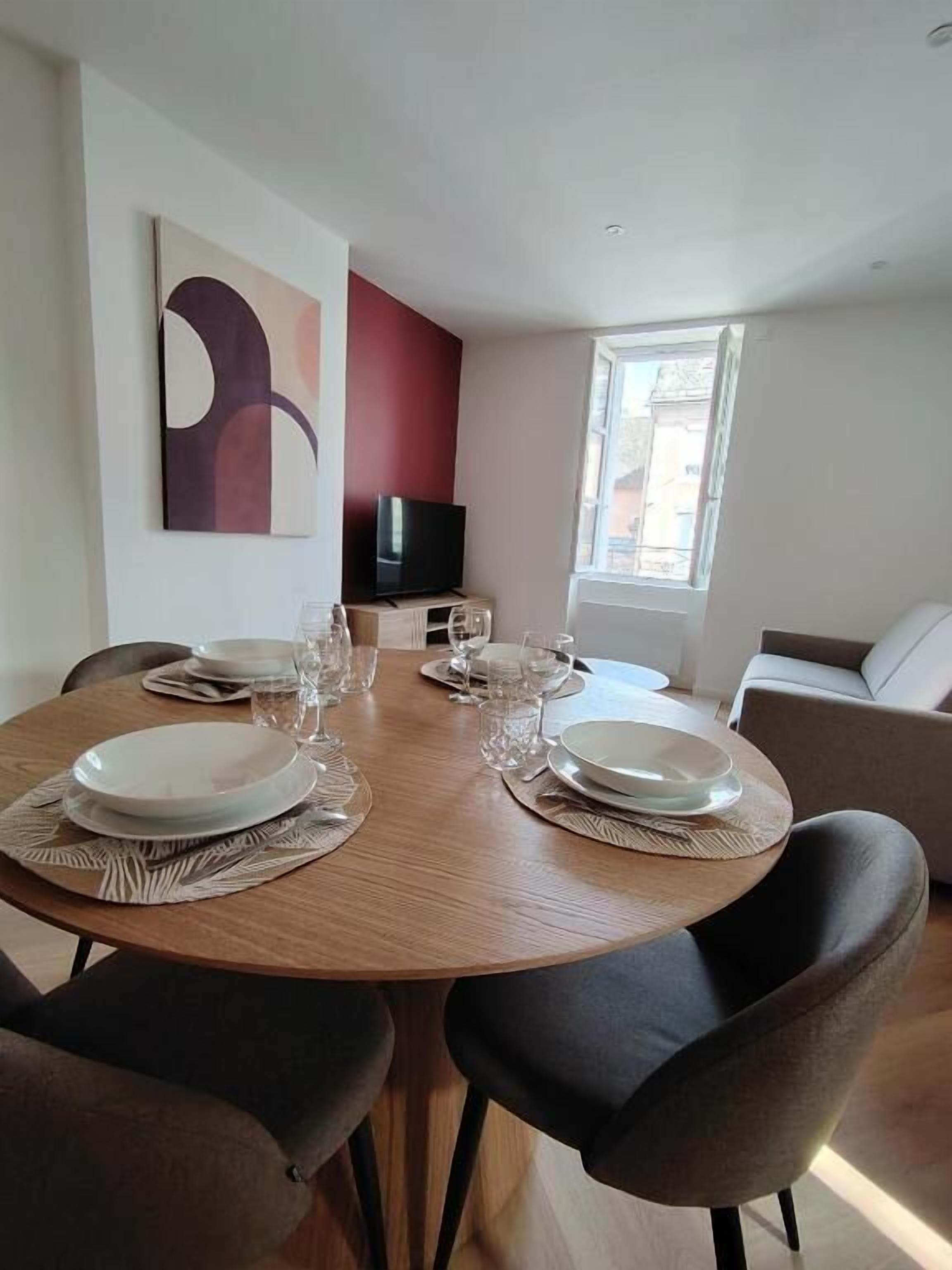 Un coin repas avec une table ronde dressée pour quatre personnes, un téléviseur posé sur un meuble et une fenêtre offrant une vue agréable.