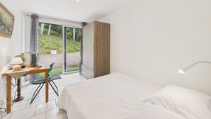 A bedroom with a bed, a desk with a lamp, a chair, and a view of greenery through a glass door. - 700 m from beach , flat for 4 with terrace (Jard-sur-Mer)