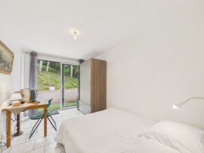 Ein Schlafzimmer mit Bett, Schreibtisch mit Lampe, Stuhl und Blick auf Grünanlagen durch eine Glastür.