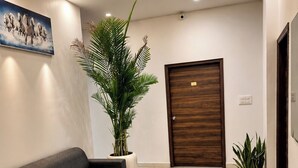 Een moderne lobby met een bank, een salontafel met glazen blad, potplanten, een houten deur en een schilderij aan de muur.