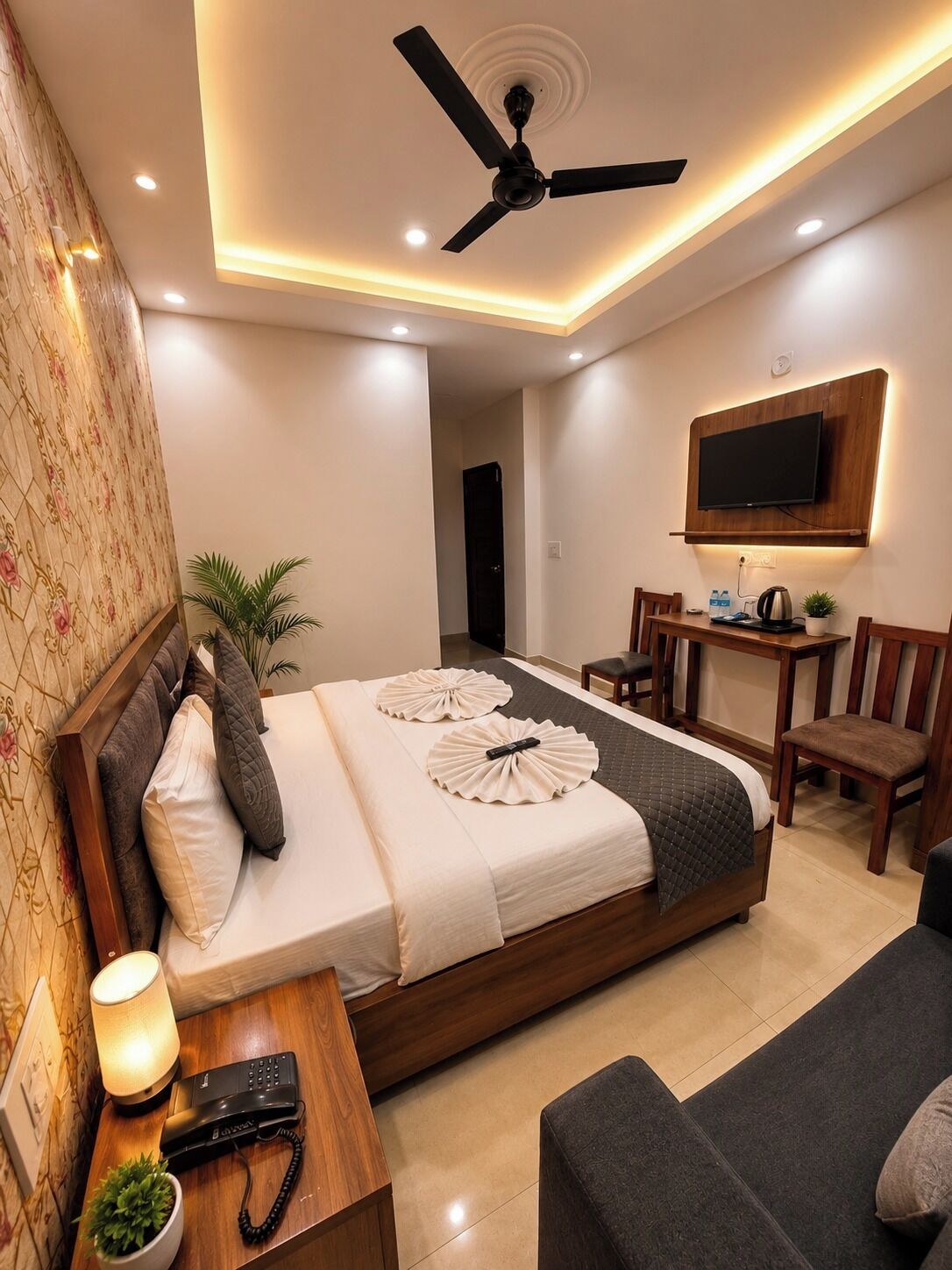 A hotel room with a bed, bedside table with a lamp, a desk with a chair, a TV mounted on the wall, and a ceiling fan.