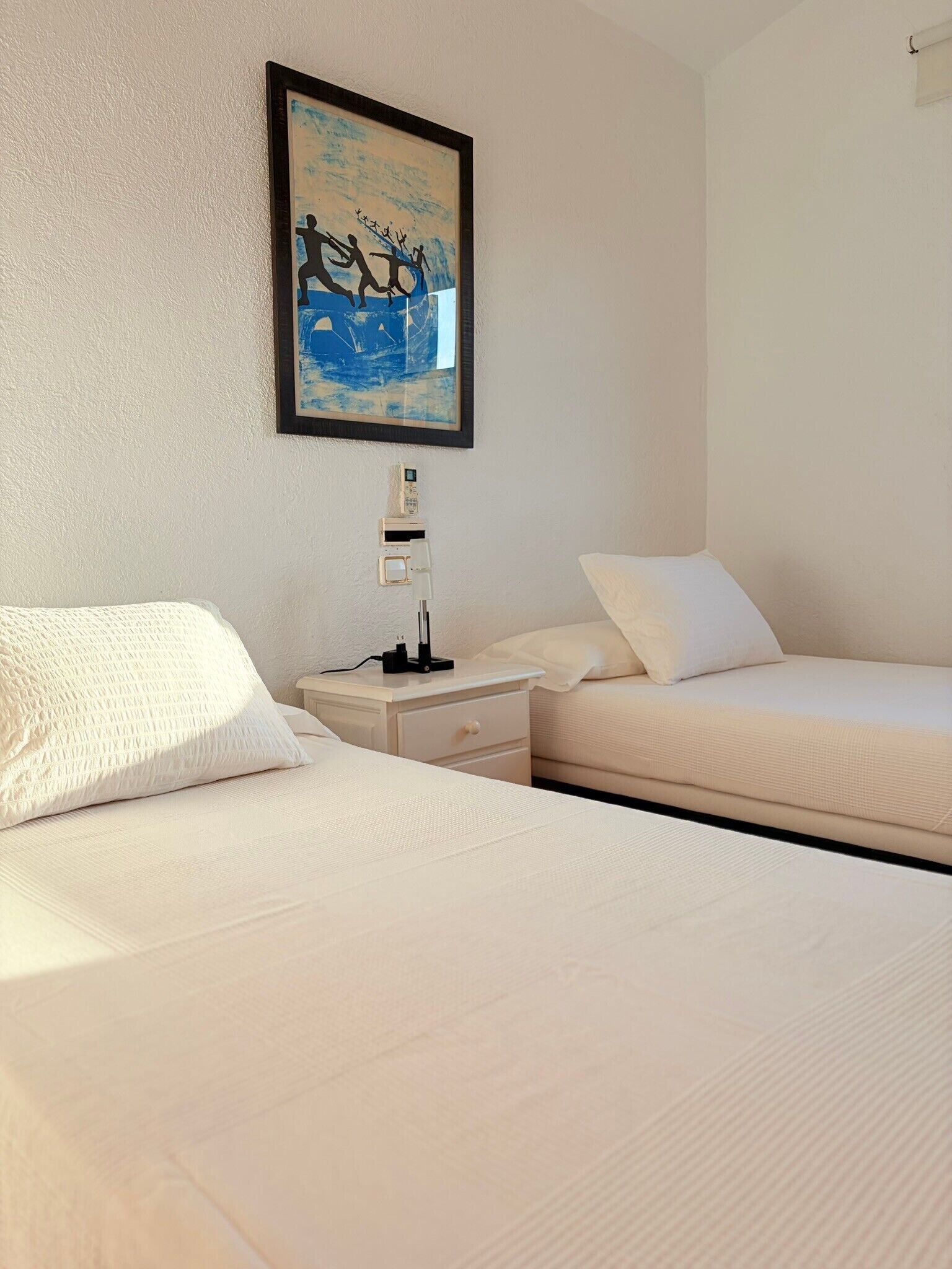 A hotel room with two beds, a nightstand with a lamp, a painting on the wall, and a power outlet.