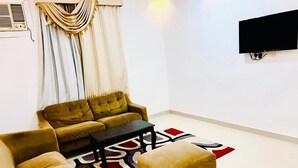A living room with a sofa, ottoman, and coffee table, a patterned rug, curtains, and a mounted TV. - CROWN FURNISHED APARTMENTS SALALAH (SALALAH)