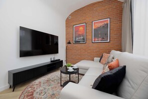 Ein Wohnzimmer mit Flachbildfernseher, einer schwarzen Unterhaltungsanlage, einem weißen Sofa, einem gemusterten Teppich und zwei gerahmten Postern an einer Ziegelwand.
