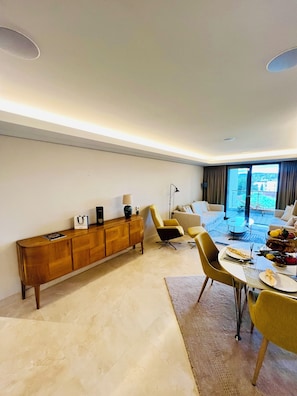 Ein modernes Wohnzimmer mit einer Holzkonsole, gelben Sessel, einem Esstisch mit Tellern und Blick durch große Fenster.