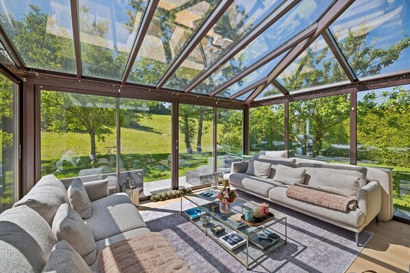 Ein modernes Wohnzimmer mit Glasdach, einer Couch, einem Couchtisch und Blick auf eine grüne Landschaft.