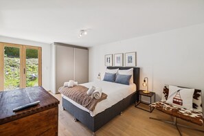 Ein Schlafzimmer mit einem Bett, Nachttischen, einer Holzkombination, einer Bank und einer Schiebetür zum Balkon.