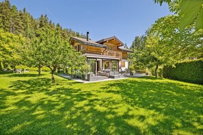 Ein zweistöckiges Holzhaus mit einer überdachten Terrasse, umgeben von einem gepflegten Rasen und Bäumen.