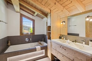 Ein Badezimmer mit Badewanne, zwei Waschbecken, einem großen Spiegel, Holzbalkendecke und einem Fenster.