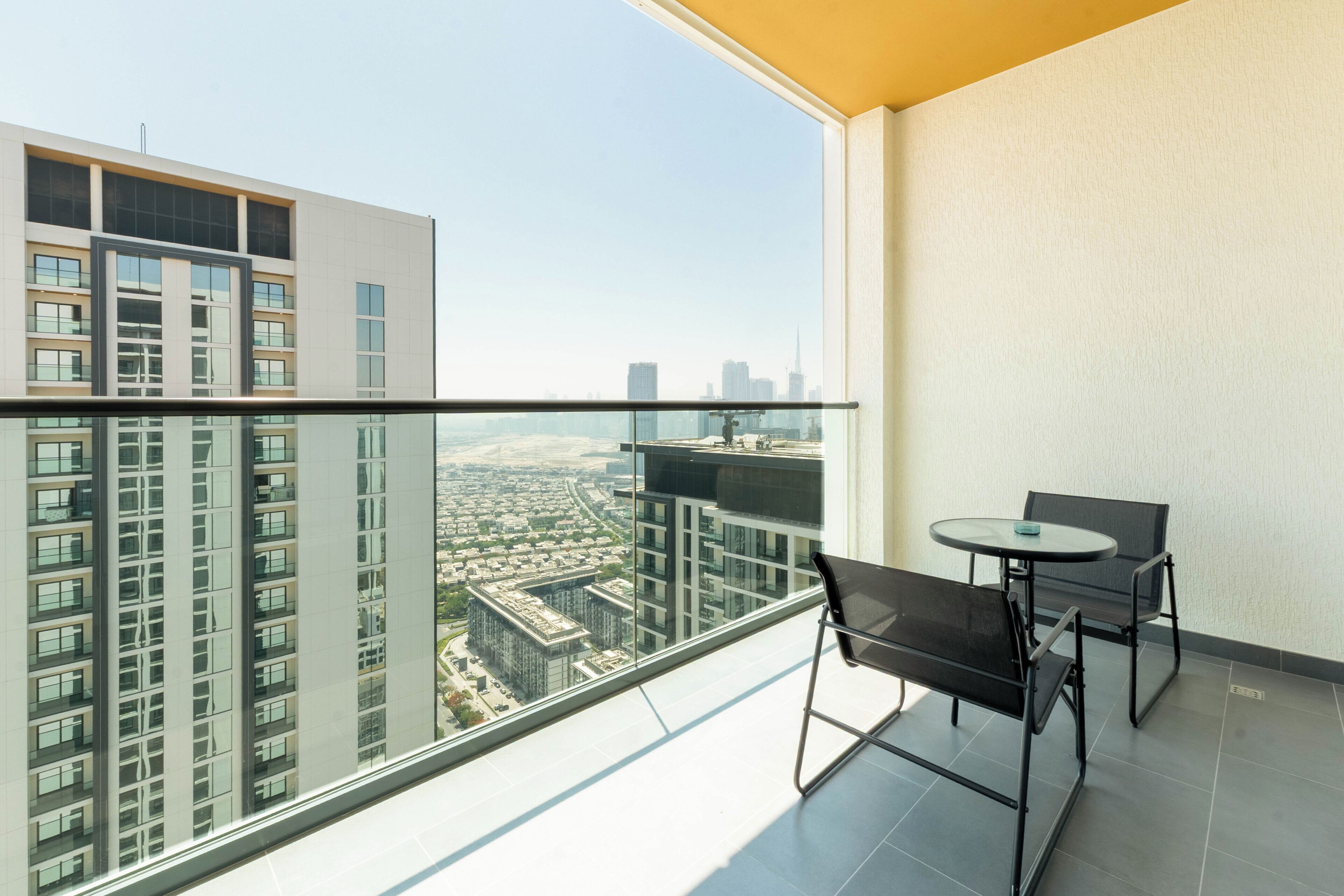 A balcony with a glass railing, a table, two chairs, and a view of a cityscape.