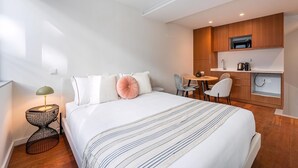 Quarto de hotel com cama, mesa de cabeceira, luminária e área de kitchenette equipada com micro-ondas e máquina de café.