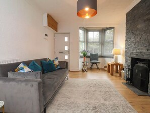 Ein Wohnzimmer mit grauem Sofa, einem kleinen Tisch mit Lampe, einem Kamin und Blick durch ein Fenster mit Jalousien.