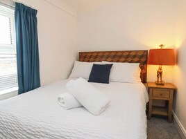 A neatly made bed with white bedding and a rolled white towel, a wooden headboard, a bedside table with a lamp, and a window with blue curtains.