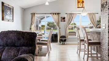 A living room with a sofa, dining table, chairs, and a view through large windows.