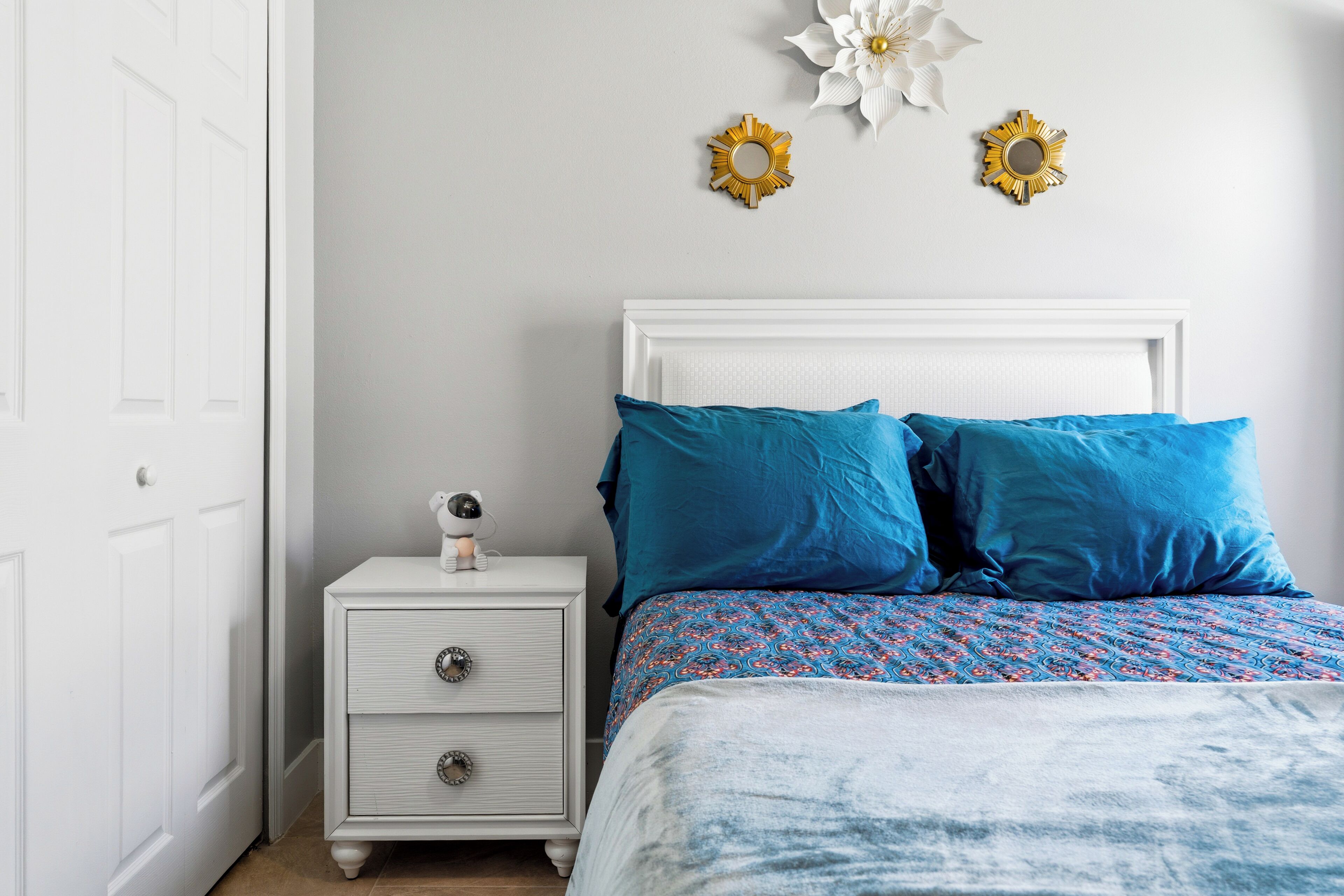A bedroom with a white bed, blue pillows, a white nightstand with drawers, and two wall decorations.