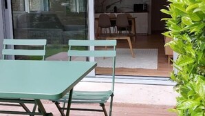 Une terrasse avec une table et des chaises, une porte vitrée donnant sur la cuisine et une plante verte sur la droite.