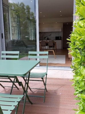 A patio with a table and chairs, a glass door leading to a kitchen, and a green plant on the right.