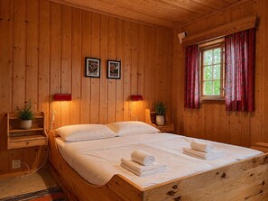 A wooden-paneled room with a bed, two framed pictures on the wall, a window with red curtains, and a small plant on a nightstand.